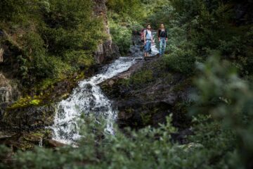 Zwei Personen stehen am Rand eines Wasserfalls mitten im Grün