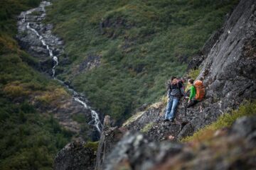 Zwei Wanderer rasten an einem Berg mit Blick auf einen gegenüberliegenden Wasserfall