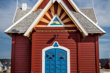 Rote Kirche mit blauer Türe und gelben Streifen am Oberlicht unter dem weißen Giebel