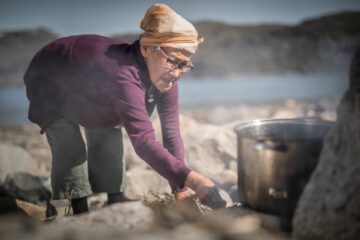 Ältere Frau mit Kopftuch bückt sich zur Feuerstelle unter Kochtopf