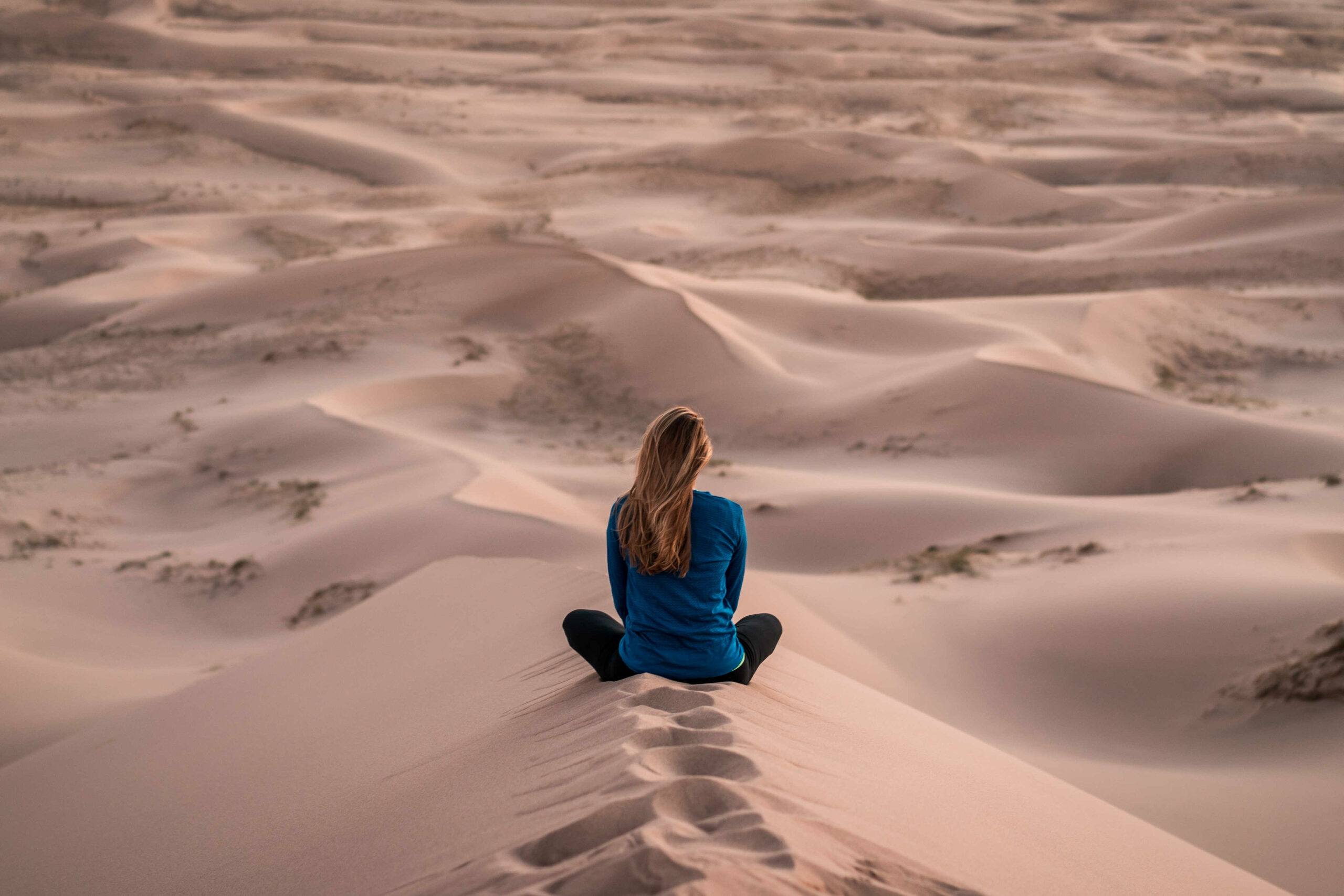 Frau in Meditationspose in einer Sanddüne – Sinnbild für Entschleunigung, Fokus und die regenerative Kraft der Stille.