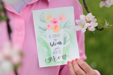 "In order to bloom, you need to grow." - Text gemalt mit Wasserfarben auf einer Karte, die von einer Frau gehalten wird.